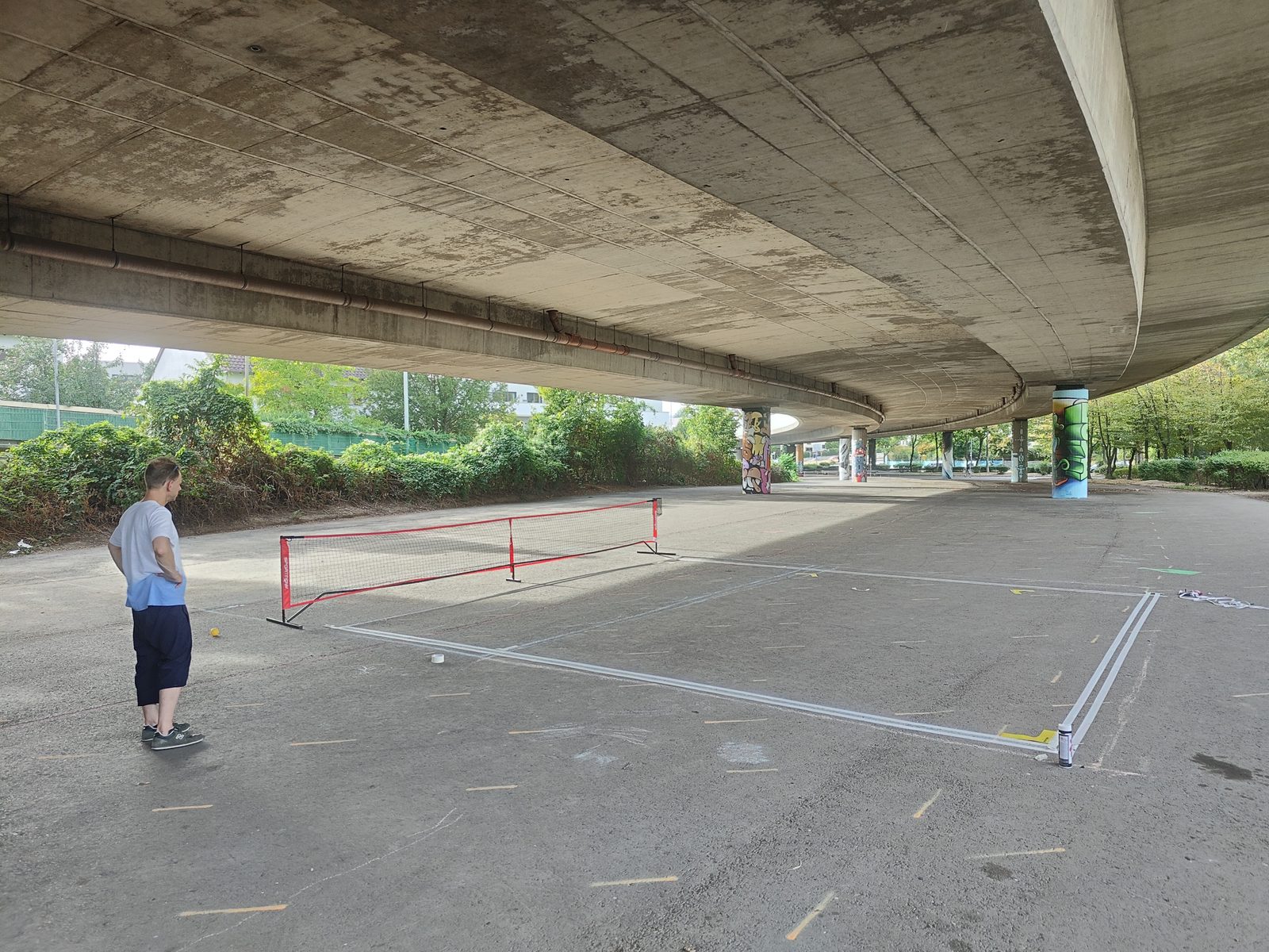 Blick über das Spielfeld unter der Brücke, eine Person steht am Rand neben dem Netz, dahinter die bemalten Brückenpfeiler.