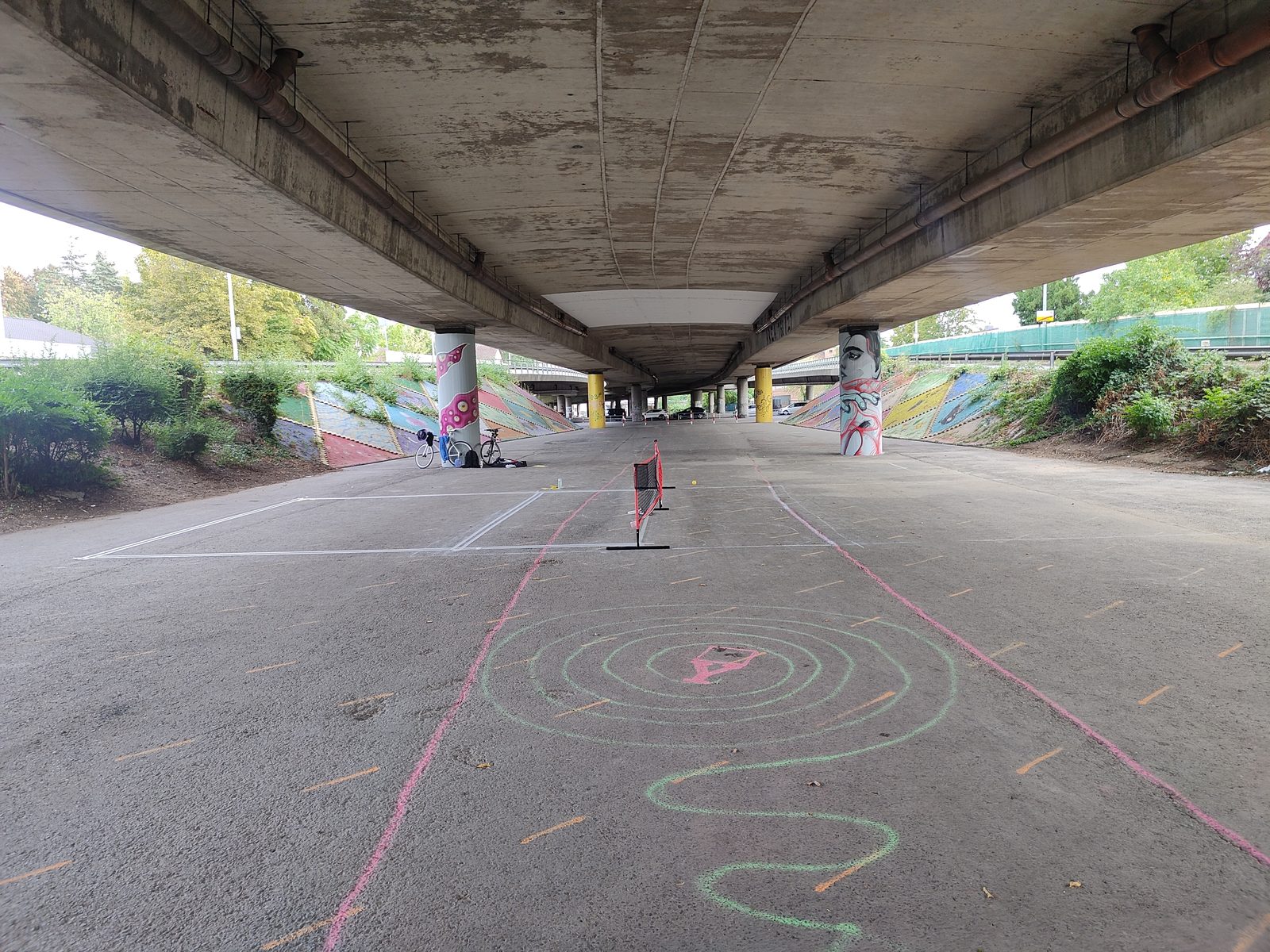 Weite Ansicht der Fläche unter dem Brückendeck mit Kreidebildern auf dem Asphalt und dem Netz im Hintergrund.